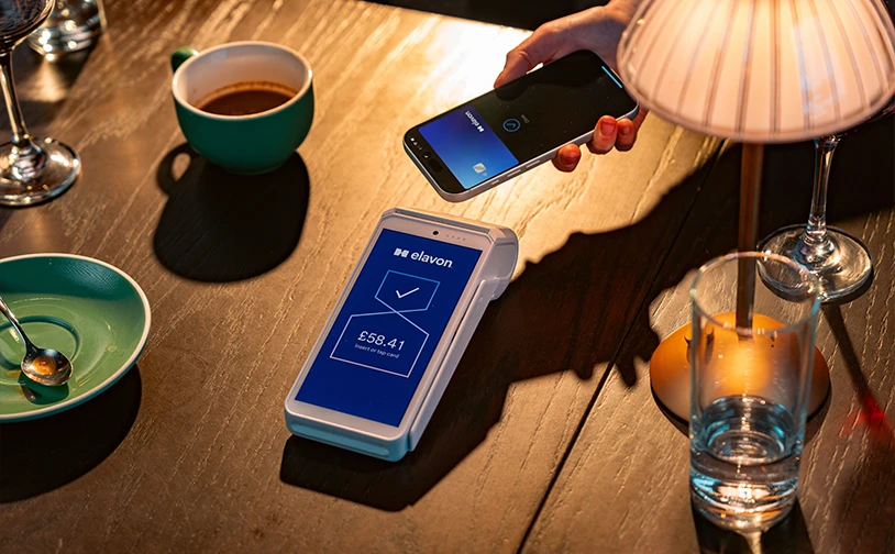A customer makes a contactless payment with a smartphone on an Elavon card machine at a restaurant table with drinks and coffee