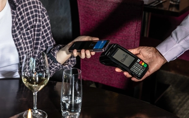 A diner in Ananda restaurant uses Apple Pay on a smartphone to make a contactless payment via an Elavon-branded terminal. The screen reads 'Hold Near Reader' as the transaction is initiated.