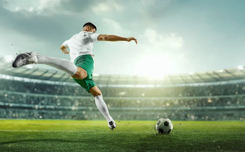 A football player in a blue uniform prepares to kick a ball on a grassy field, with a stadium full of spectators in the background under a cloudy sky