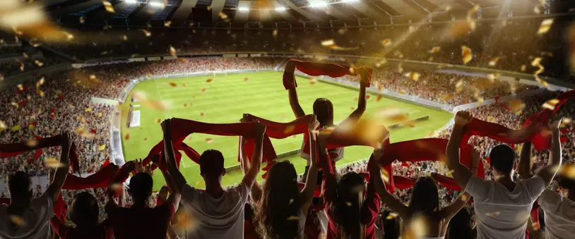 Fans holding red scarves celebrate in a packed, well-lit stadium with confetti in the air and a green field in the background.