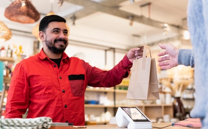Retail staff handing a takeaway shopping bag to a customer at a checkout counter with card payment terminal
