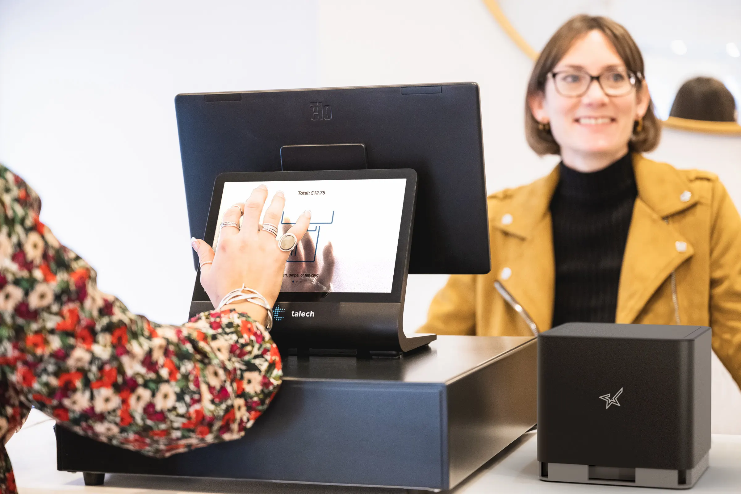 An employee at a shop checkout uses a touchscreen payment terminal, while a shopper stands on the opposite side of the counter 