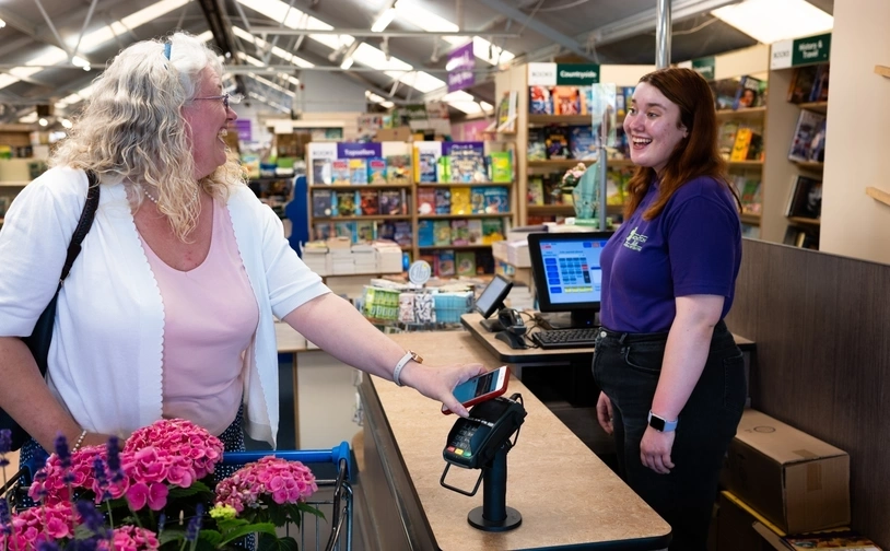 A customer at a checkout counter holds a payment card near a contactless reader while speaking with a store employee in a retail environment. Bright flowers and shelves of products are visible in the background