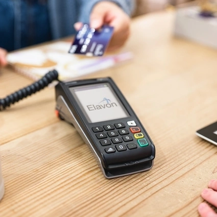 A payment terminal with the Elavon logo on the screen, sitting on a wooden counter as a customer makes a secure card payment in-store. 